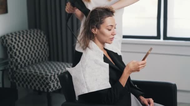 Zblízka, kadeřnice vysuší žena mokré vlasy s fénem, klient používá mobilní — Stock video