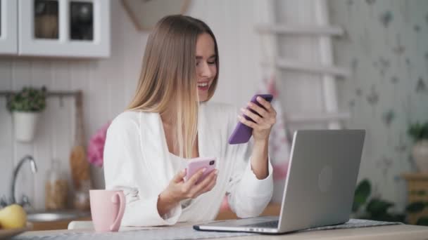 Молода жінка сидить за столом з ноутбуком на кухні, використовує два телефони — стокове відео