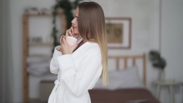 Mooie jonge vrouw in badjas dansen en praten over de telefoon in de slaapkamer — Stockvideo
