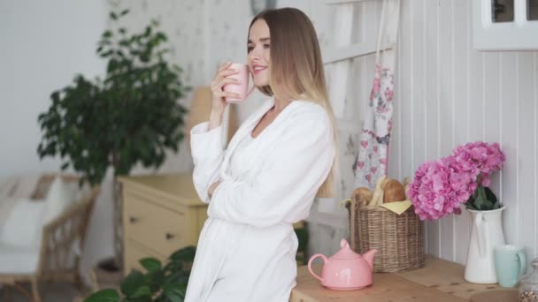 Fiatal nő fürdőköpenyben iszik teát egyedül a konyhában, élvezi a reggeli órákat, álmokat — Stock videók