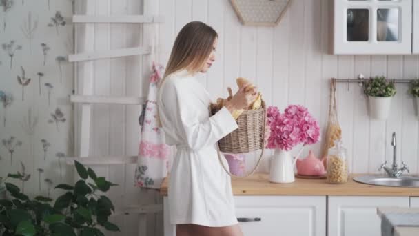 Mujer joven en albornoz en la cocina toma cesta de pan y elige más fresco — Vídeo de stock