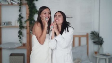 Beyaz bornozlu iki kız arkadaş dans eder, şarkı söyler, saç kurutma makinesiyle saçlarını kurutur, güler.