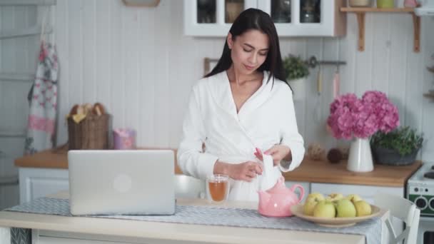 Młoda kobieta w białym szlafroku pracuje na laptopie i pije herbatę rano — Wideo stockowe