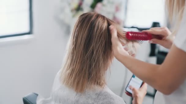 美容師の手を閉じると、女性のぬれた髪を結合し、クライアントはスマートフォンを使用しています — ストック動画