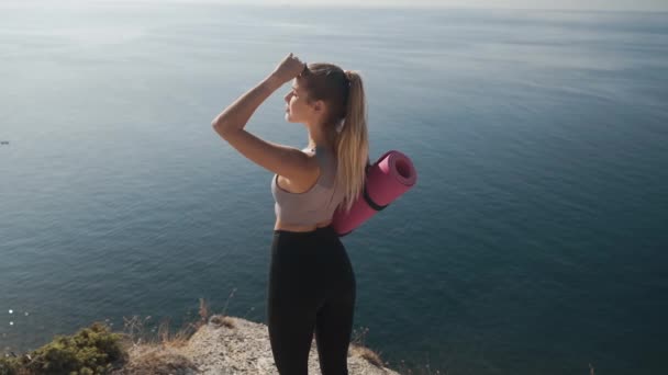 Femme tient tapis de yoga, regarde la mer de falaise, respire profondément, au ralenti — Video