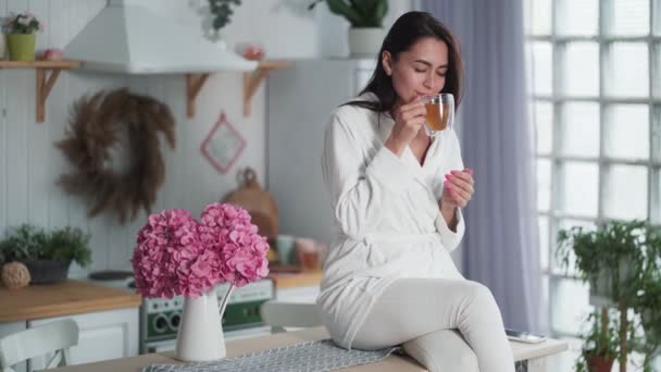 Glückliche Frau sitzt am Küchentisch, trinkt heißen Tee, genießt den Morgen, träumt — Stockvideo