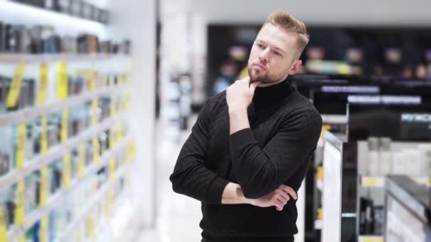 Homem barbudo pensivo olha prateleiras com mercadorias e escolhe o que comprar no shopping — Vídeo de Stock