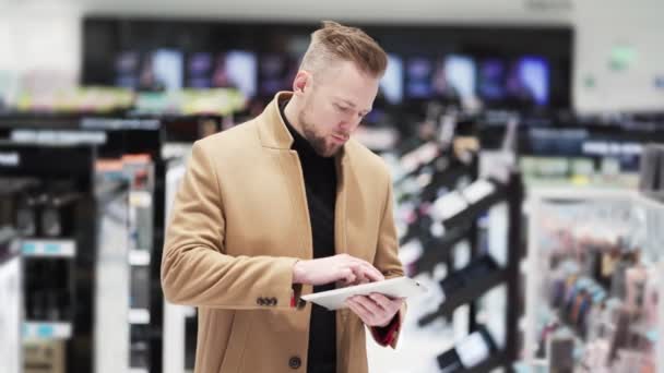Jonge knappe man met baard in bruine jas gebruikt digitale tablet in het winkelcentrum. — Stockvideo