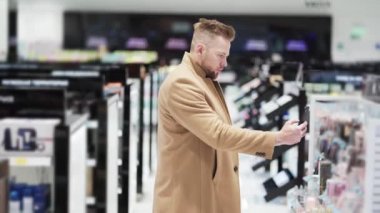 Kahverengi ceketli genç adam telefon kullanır ve kozmetik mağazasında malların fotoğrafını çeker.