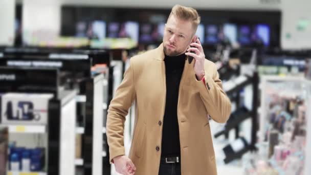 Young handsome bearded man in brown coat talking on phone in shopping mall. — Stock Video