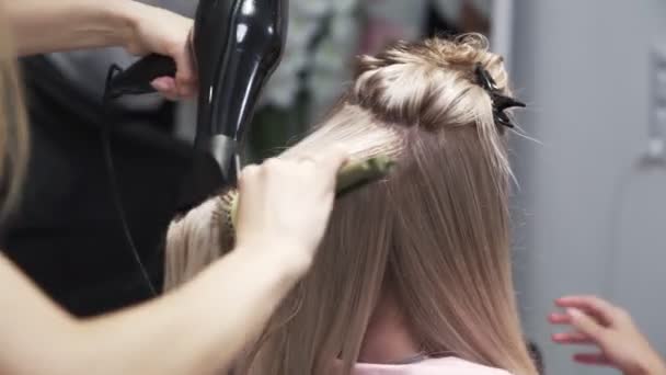 Yaklaş, kuaför uzun sarı kadın saçını saç kurutma makinesiyle kurutur ve saçlarını yuvarlak tarar. — Stok video
