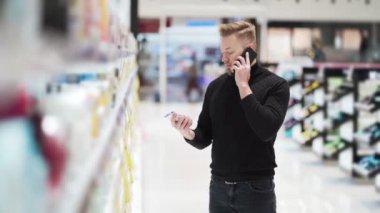 Genç sakallı adam telefonla konuşuyor ve kozmetik dükkanından ürün seçiyor.
