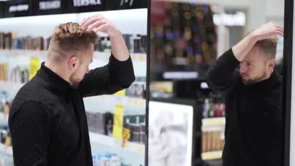 Jonge man met baard kijkt naar spiegel in cosmetica winkel en raakt zijn haar — Stockvideo