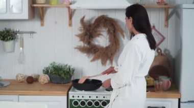 Beyaz bornozlu genç bir kadın mutfakta yemek pişiriyor.