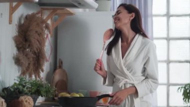Beyaz bornozlu genç bir kadın mutfakta yemek pişiriyor ve evde dans ediyor.