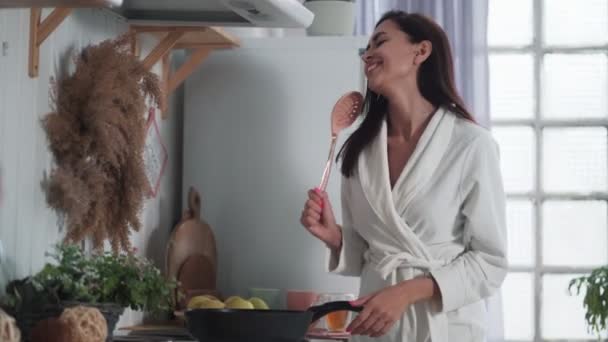 穿着白色浴衣的年轻女子在厨房做饭，在家里跳舞 — 图库视频影像