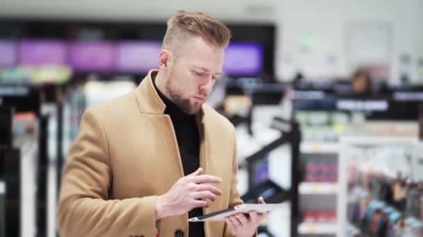 Jonge knappe man met baard in bruine jas gebruikt digitale tablet in het winkelcentrum. — Stockvideo