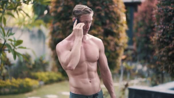 Shirtless man with muscular body talking on phone outdoor, greens on background — Stock Video