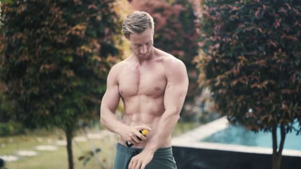 Sportovní sexy muž aplikuje opalovací olej na jeho tělo s bazénem na pozadí — Stock video