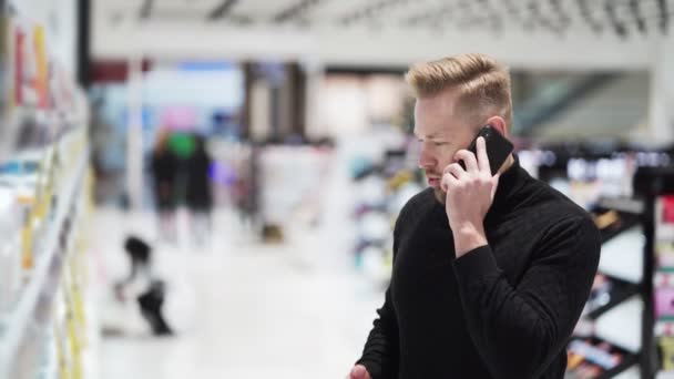 Jonge man met baard praten over de telefoon en selecteert product in cosmetica winkel — Stockvideo