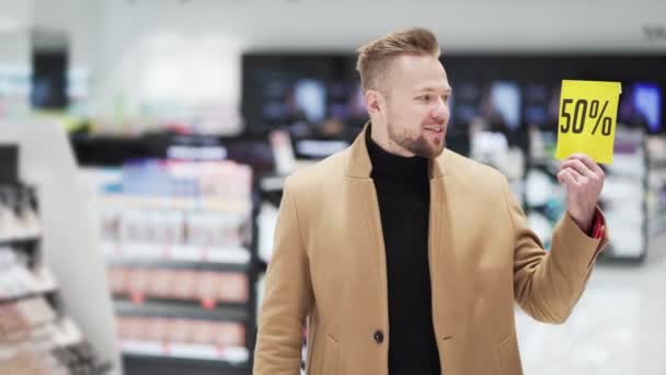 Молодой бородач в магазине косметики показывает два плаката со скидкой с большой распродажей — стоковое видео