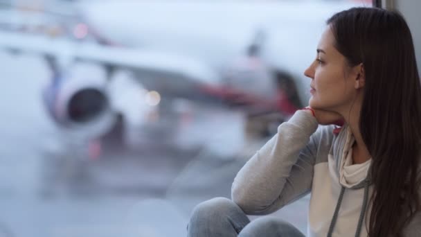 Jonge vrouw zit op het vliegveld in de buurt van venster, wazig vliegtuig op de achtergrond — Stockvideo