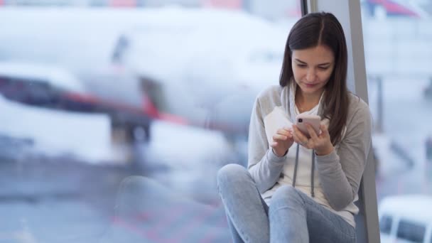 Vrouw zit op de luchthaven en maakt gebruik van telefoon, wazig vliegtuig op de achtergrond — Stockvideo