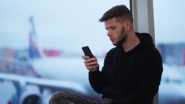 Jovem sentado no aeroporto e usa telefone, avião embaçado no fundo — Vídeo de Stock