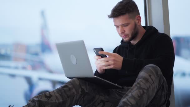 Hombre sentado en el aeropuerto y utiliza teléfono inteligente y portátil, avión en el fondo — Vídeos de Stock