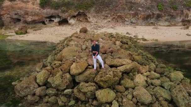 Aerial view of a man in white pants and a shirt stands on stones near the ocean — 비디오