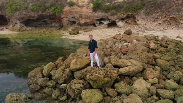 Aerial view of a man in white pants and a shirt stands on stones near the ocean — 비디오