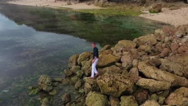 Aerial view of a man in white pants and a shirt stands on stones near the ocean — Stock Video