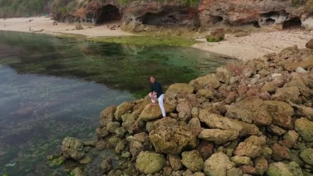 Aerial view of a man in white pants and a shirt stands on stones near the ocean — Stock Video