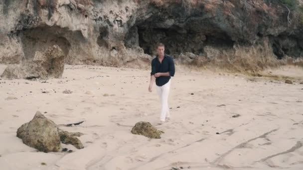 Samotny mężczyzna w białych spodniach i czarnej koszuli spaceruje wzdłuż piaszczystej plaży. — Wideo stockowe