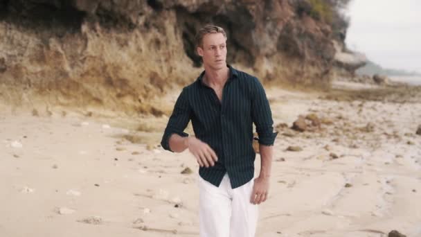 Magányos férfi fehér nadrágban és fekete ingben sétál a homokos parton.. — Stock videók