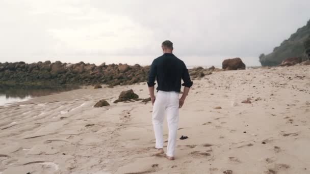 Lonely man in white pants and shirt walks along the sandy beach, backside view. — 비디오