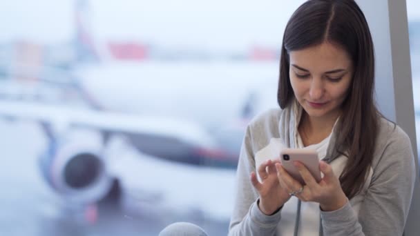 Vrouw zit op de luchthaven en maakt gebruik van telefoon, wazig vliegtuig op de achtergrond — Stockvideo