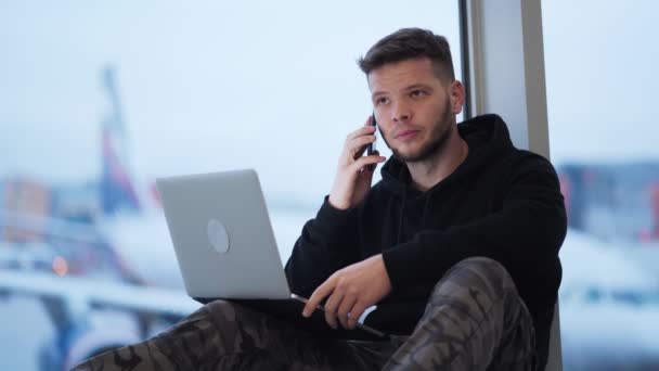 Чоловік сидить в аеропорту і використовує смартфон і ноутбук, літак на фоні — стокове відео