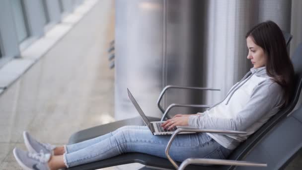 Mujer joven se sienta en la sala de salida y utiliza el ordenador portátil para el trabajo en el aeropuerto — Vídeos de Stock