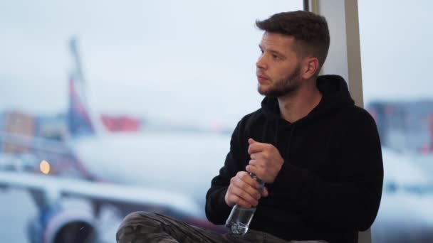 Man drinkt water uit plastic fles op de luchthaven, wazig vliegtuig op de achtergrond — Stockvideo