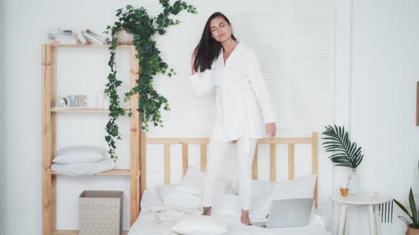 Feliz joven en albornoz blanco saltando y bailando en la cama con almohadas — Vídeos de Stock