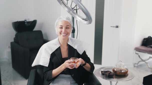 Güzellik salonunda saç bakımı yapan genç bir kadın, çay içiyor ve rahatlıyor. — Stok video