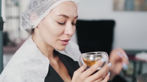 Hermosa mujer en la tapa cosmética bebe té en el salón de belleza, procedimiento de cuidado del cabello — Vídeos de Stock