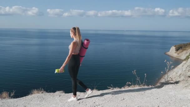 Mujer joven en ropa deportiva con colchoneta de gimnasia se encuentra en el borde del acantilado — Vídeos de Stock