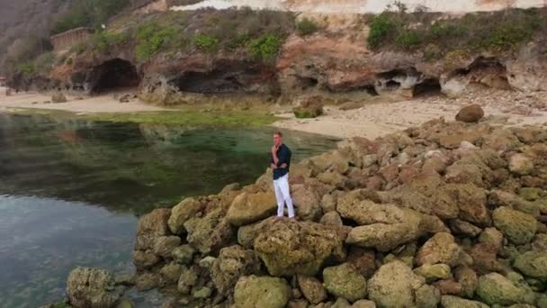 Légi felvétel egy fehér nadrágos férfiról és egy ingről az óceán melletti köveken áll. — Stock videók
