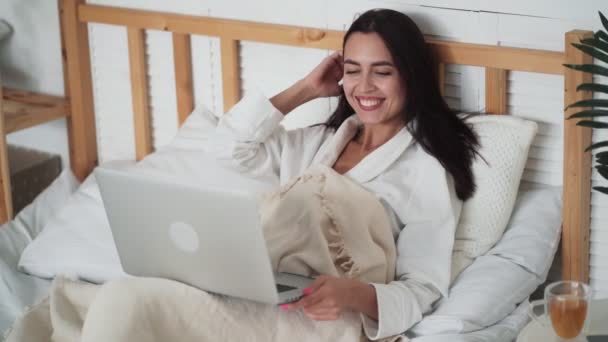 Portret van jonge vrouw in badjas ligt in bed en heeft video chat op laptop — Stockvideo