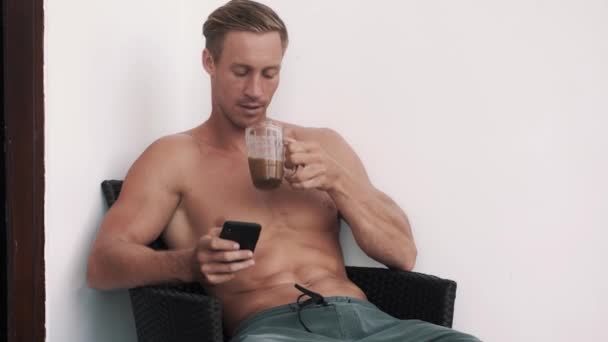 Sexy muž bez trička sedí v křesle, pomocí telefonu a pití kávy venku — Stock video