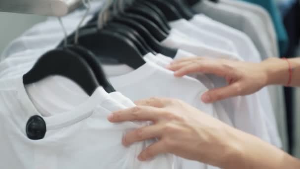 Primo piano delle mani della donna, guardare attraverso i vestiti su rack, acquirente sceglie t-shirt bianca — Video Stock