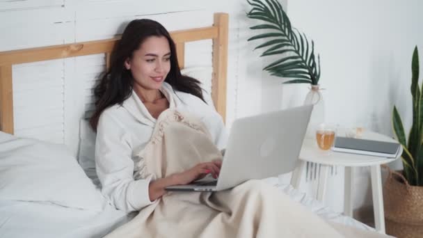 Hermosa mujer joven se encuentra en la cama, utiliza el ordenador portátil para el trabajo o chat en línea — Vídeos de Stock