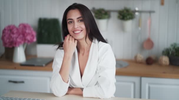 Портрет милой молодой женщины сидит дома на кухне, смотрит в камеру, улыбается — стоковое видео